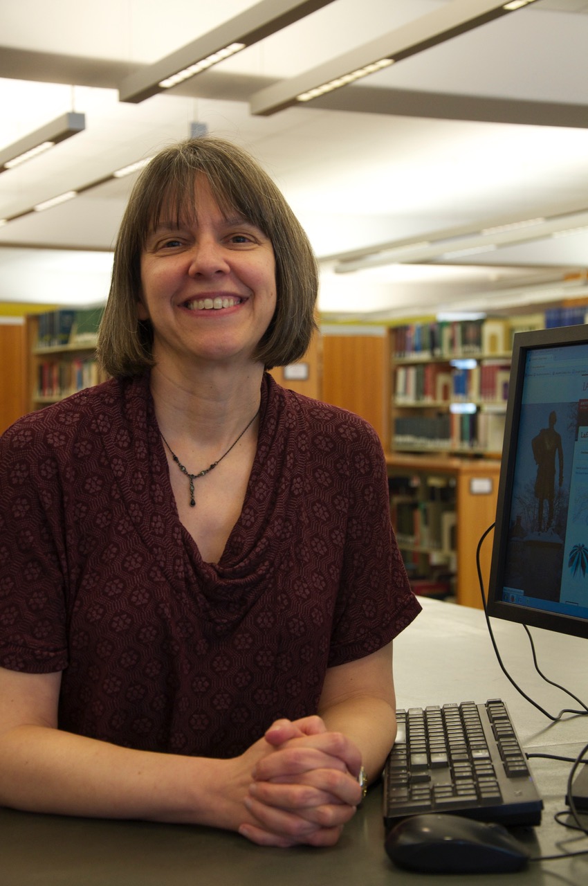 Terese Heidenwolf sits at the reference desk in front of a computer. Her hands are folded and a large smile is on her face.