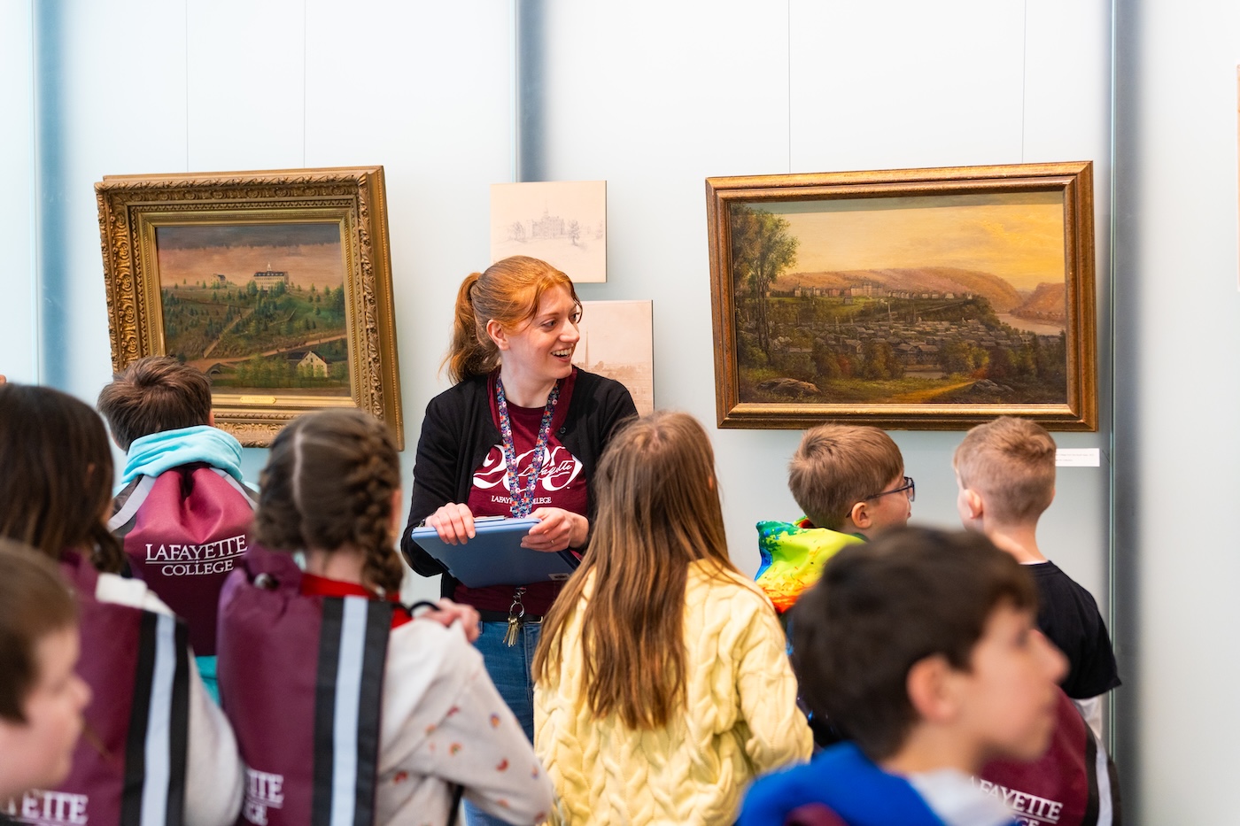Missy Nerino stands in front of an art display wall, surrounded by children looking at her. She holds a clipboard.